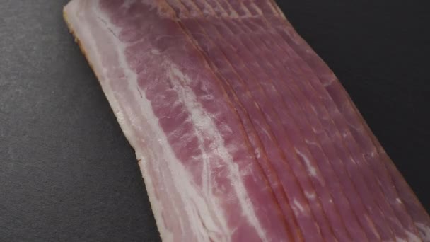 Hand takes a strip of fresh red bacon with fat layers. On a black stone board. Sliced appetizing meat - Footage, Video