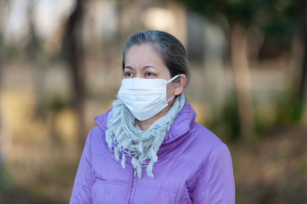 Gyönyörű kevert fajú középkorú nő fehér maszkot visel a koronavírus (COVID-19) és más fertőző betegségek elleni védelemre. Félrenézek. Homályos kültéri háttér.  - Fotó, kép