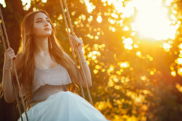 beautiful girl with long hair swinging on rope swing on summer nature ar sunset, young woman enjoy flying among tree branches, leisure activity, lifestyle concept - 写真・画像