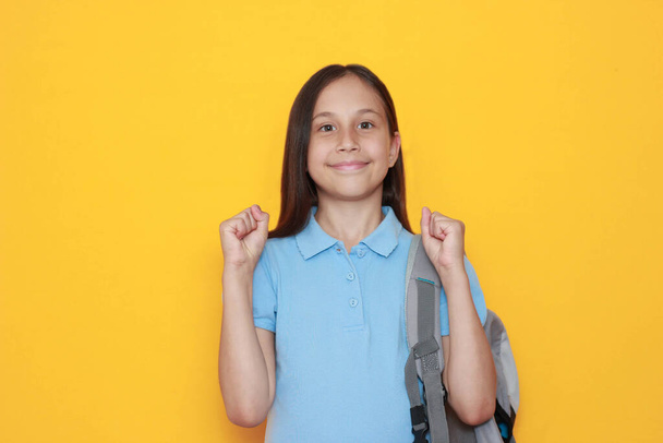a cute smiling brunette girl schoolgirl with a backpack on her shoulder with bent arms at the elbows shows cool, cool, yes on a yellow background - Photo, Image