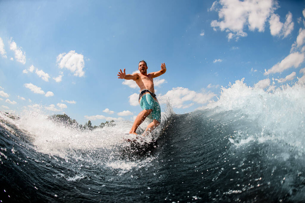 эмоциональный взрослый парень активно катается по волнам на доске для серфинга против голубого неба - Фото, изображение
