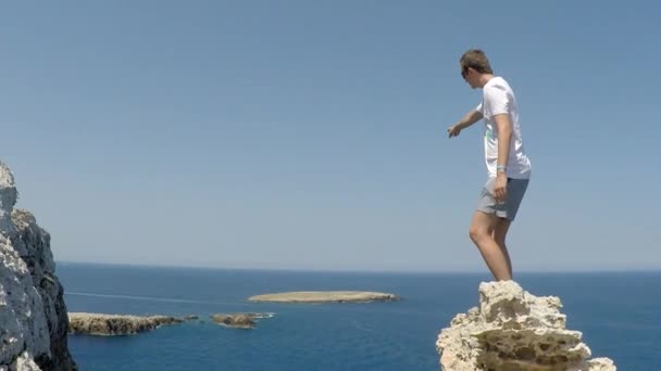 Junger Reisender hebt die Hände hoch oben auf dem Berg über der wunderschönen Landschaft. Ein junger Mann auf der Klippe des Cap Cavaleria starrt aufs Meer und zeigt mit der Hand weg, Menorca, Spanien. - Filmmaterial, Video