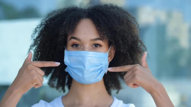 Beautiful smiling young African woman shows with two fingers wearing mask to protect against outbreak of coronavirus during second wave of pandemic. Ethnic girl student showing thumbs up, close up - Πλάνα, βίντεο