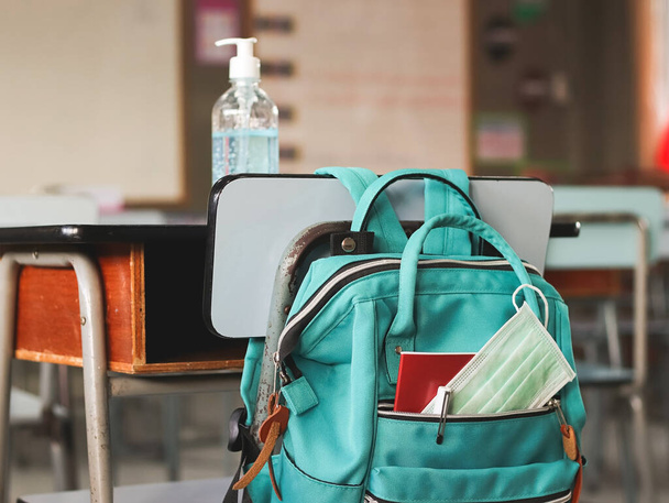 COVID-19 önleme, okula geri dönüş ve yeni normal kavram. Okul malzemeleriyle dolu okul çantası ve sınıfta masasında alkol jeliyle sallanan sandalyeli sağlık maskesi.. - Fotoğraf, Görsel