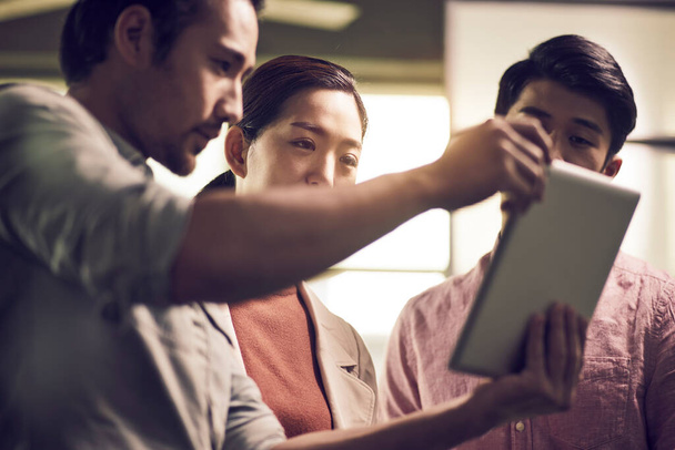Drei junge asiatische Unternehmer arbeiten im Büro zusammen und schauen auf ein digitales Tablet - Foto, Bild