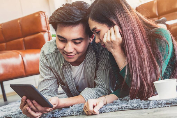 Feliz pareja asiática utilizar la tableta mientras está acostado en la alfombra en el piso de la sala de estar. Concepto de amor y relación
. - Foto, imagen
