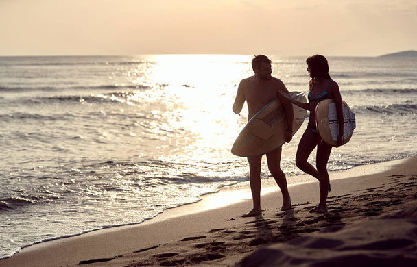 young couple walking on the sandy beach at sunset, carrying surfboards, talking - Photo, image