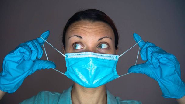 Μια γυναίκα γιατρός ή νοσοκόμα με λαστιχένια γάντια στα χέρια βάζει μια μπλε μάσκα στο πρόσωπό της. - Φωτογραφία, εικόνα