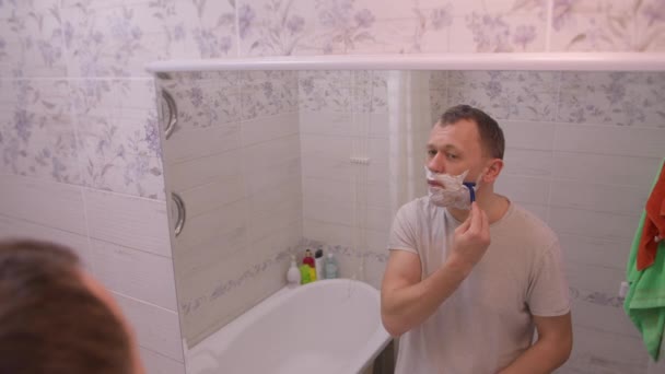 Een man scheert, kijkt naar zichzelf in de spiegel, achteraanzicht, camerabeweging - Video