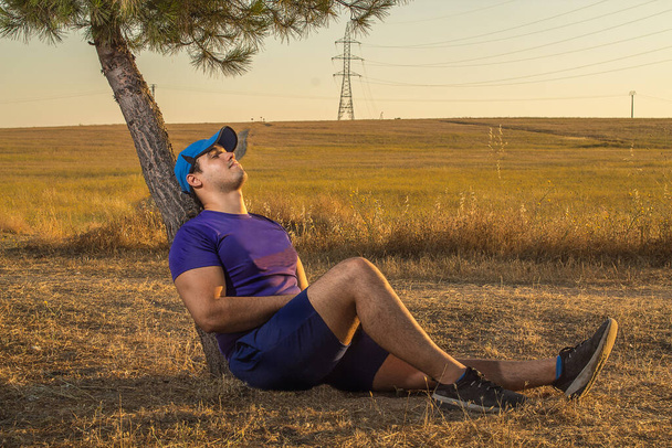 Кавказький молодий чоловік зі спортивними фіолетовими сорочками і ковпаком на дереві після бігу.. - Фото, зображення