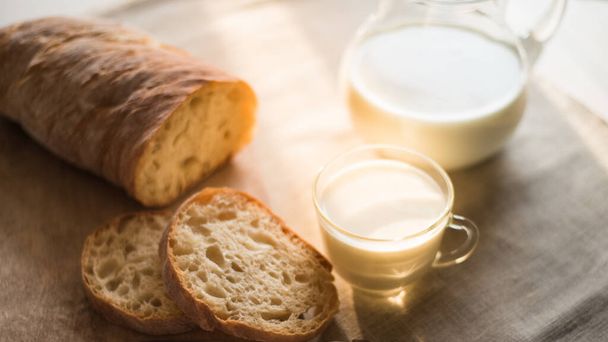Light rustic breakfast - rustic bread with cut pieces, a jug and a glass of milk on a linen napkin on the table by the window, morning light, panoramic view - Photo, Image