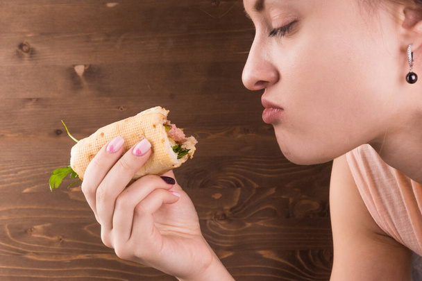 Mladý atraktivní žena jíst hot dog na pozadí hnědé dřevěné zdi, close-up - Fotografie, Obrázek