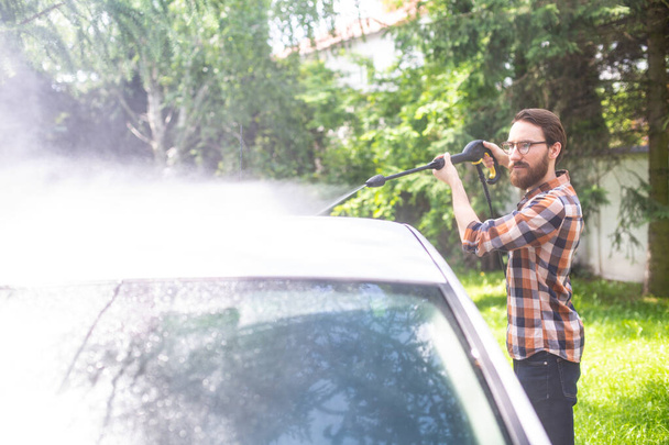 Car washing in the garden with a high pressure washer on a warm sunny day. A young bearded man using a compact home high pressure washer. Backyard, exterior, outdoor car cleaning. - Fotó, kép
