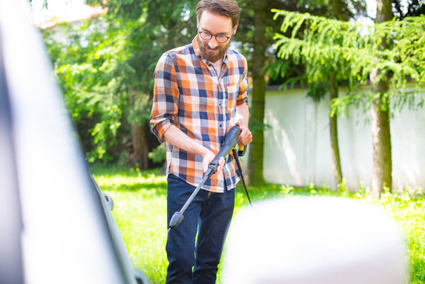 Car washing in the garden with a high pressure washer on a warm sunny day. A young bearded man using a compact home high pressure washer. Backyard, exterior, outdoor car cleaning. - Φωτογραφία, εικόνα