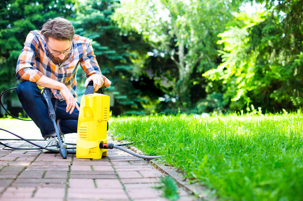 A young man works in the garden using a yellow compact home high pressure washer. Garden, patio, backyard cleaning on warm sunny day. An outdoor pressure washing, greenworks, cleaning car and exterior - Fotografie, Obrázek