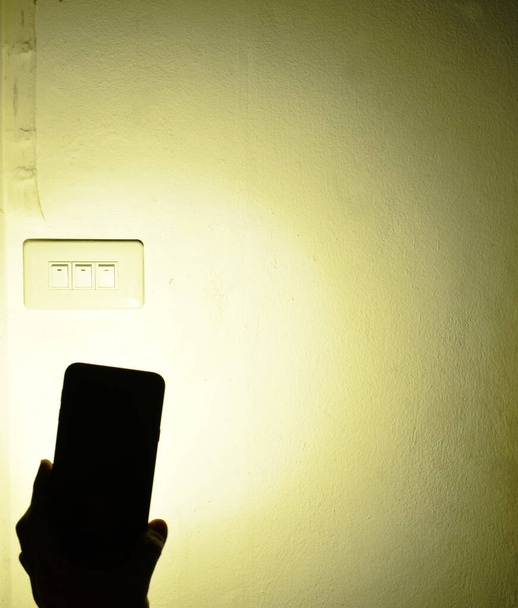 ліхтарик мобільного телефону, що поширює світло на електричний перемикач на домашній стіні під час відключення електроенергії в темну ніч
  - Фото, зображення