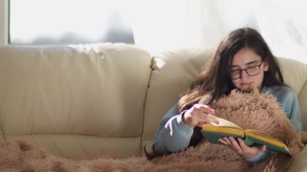 Μελαχρινή κοπέλα με γυαλιά κάθεται σε έναν καναπέ διαβάζοντας ένα βιβλίο, γυρίζοντας πάνω από σελίδες, αντίγραφο χώρο - Πλάνα, βίντεο