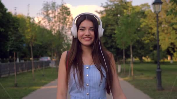 Enchanting lady is walking outdoors in park listening to music in her headphones. - Materiał filmowy, wideo