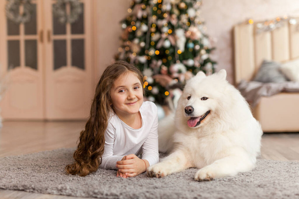Joululapsi tyttö koiran kanssa Samoyed makaa lattialla edessä suuri joulukuusi ja katselee kameraa. Joulun, talven ja ihmisten käsite. Joulutervehdyskortti. Uusi vuosi kotona - Valokuva, kuva