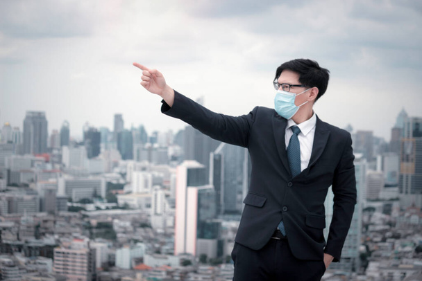 Jovem empresário asiático de terno usando máscara facial para se proteger contra vírus ou problema de poluição contra poeira de ar na cidade, homem apontando para o céu com a cidade arranha-céu
 - Foto, Imagem