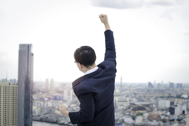 Cheerful and happy achievement successful young Asian businessman in suit raising his arm up to celebrate success goal on the rooftop with skyscraper city view - Photo, Image