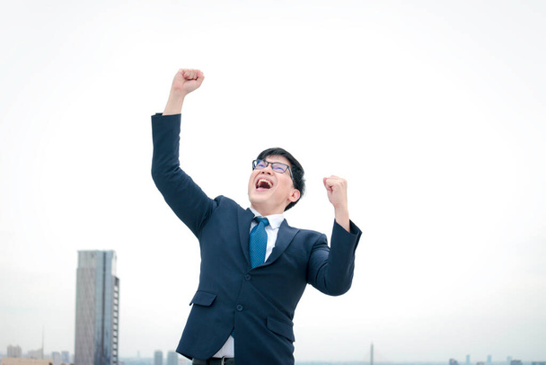 Realização alegre e feliz bem sucedido jovem empresário asiático em terno levantando o braço para celebrar o objetivo de sucesso no telhado com vista para a cidade arranha-céu
 - Foto, Imagem