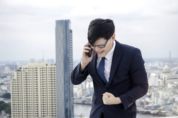 Vidám és boldog eredmény sikeres fiatal ázsiai üzletember öltönyben beszél mobiltelefon, okos telefon, felemeli a karját, hogy megünnepeljük a siker cél a tetőn felhőkarcoló városnéző - Fotó, kép