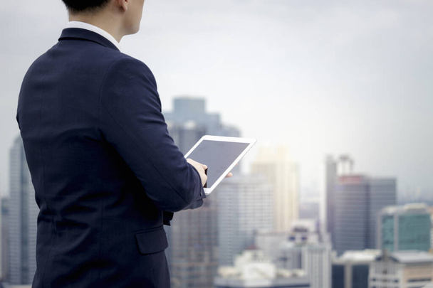 Young Asian businessman in suit using digital tablet on the rooftop with skyscraper city view - Valokuva, kuva