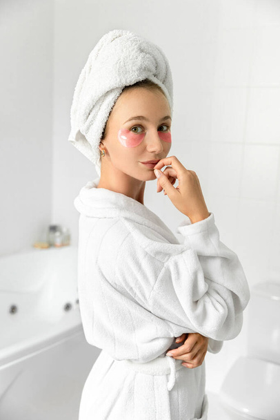 Blond schattige jonge vrouw met roze ooglapjes in witte badkamer. Ochtend wakker worden zelfverzorging thuis - Foto, afbeelding