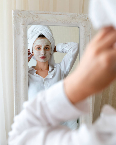 Jonge mooie vrouw aan te brengen gezichtsmasker in de slaapkamer. Spiegelreflectie. Huidverzorging routine lichaamsverzorging concept - Foto, afbeelding