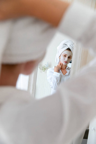 Genç ve güzel bir kadın yatak odasında yüz maskesi takıyor. Ayna yansıması. Cilt bakımı rutin vücut bakımı kavramı - Fotoğraf, Görsel