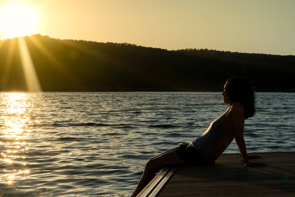 junges Mädchen mit lockigem Haar sitzt auf einer Seebrücke und blickt zum Horizont in einem Sonnenuntergang - Foto, Bild