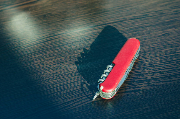 Rotes faltbares Multifunktionsmesser auf einem hölzernen braunen Hintergrund. Die Sonne scheint auf ein Messer. - Foto, Bild
