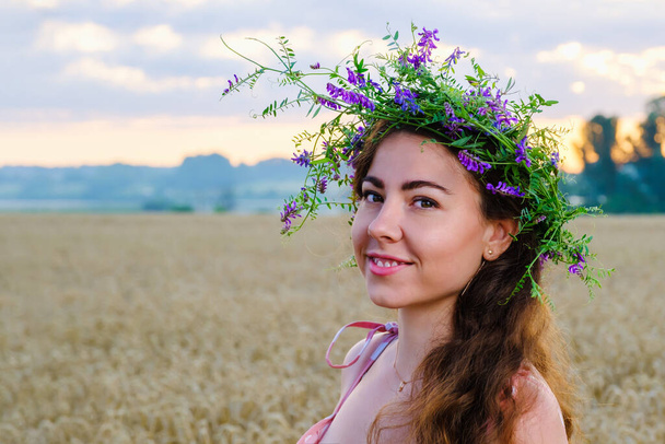 Ευτυχισμένο χαμογελαστό κορίτσι με μακριά μαλλιά με στεφάνι από λουλούδια στο κεφάλι στο χωράφι με το σιτάρι την ανατολή. Αντιγραφή χώρου - Φωτογραφία, εικόνα