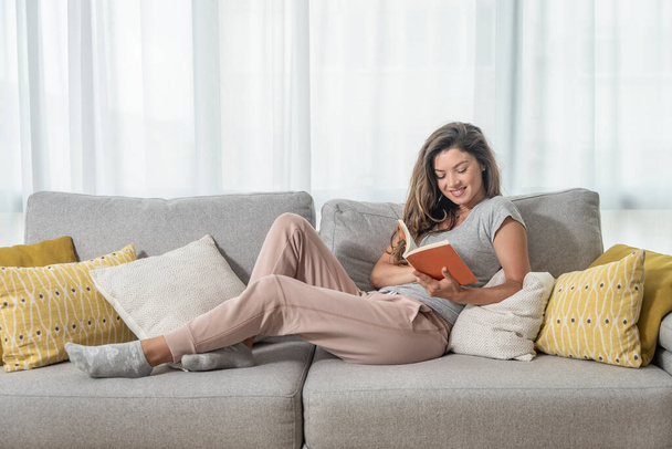 Young beautiful girl or woman lying on the sofa at home reading the book and enjoying her free time without her husband and children with smile and happy face - Valokuva, kuva