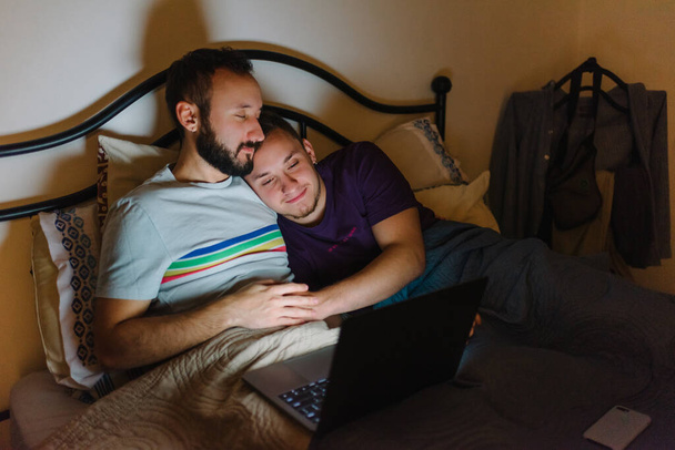 Foto de stock de dos hombres homosexuales caucásicos sentados en la cama. Están viendo algo en el portátil. Se están acurrucando
. - Foto, imagen