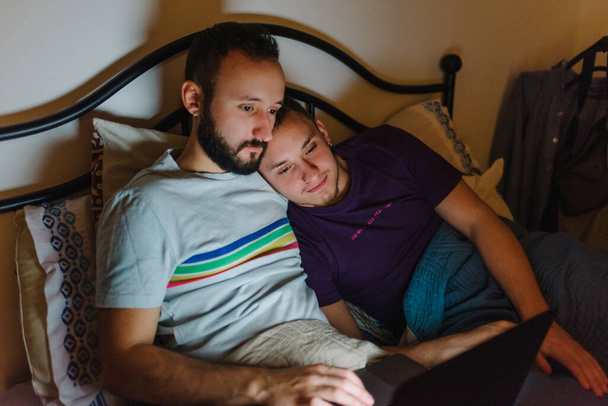 ベッドに座っている2人の同性愛者の白人男性のストック写真。彼らはラップトップで何かを見ています。抱っこしてる. - 写真・画像