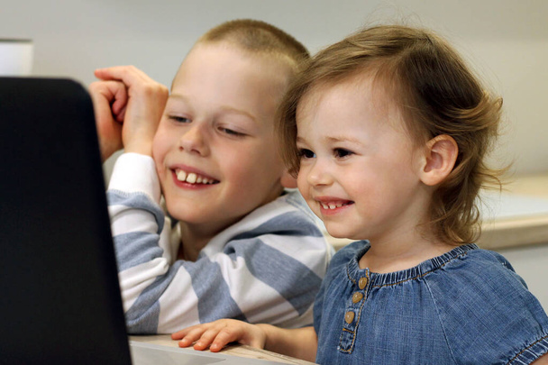 Δύο γελαστά παιδιά κάθονται στην κουζίνα και κοιτάζουν τον υπολογιστή. Τα παιδιά κοιτάζουν ένα φορητό υπολογιστή και επικοινωνούν με τους συγγενείς τους διαδικτυακά μέσω του Διαδικτύου. Έννοια της αυτοαπομόνωσης. - Φωτογραφία, εικόνα