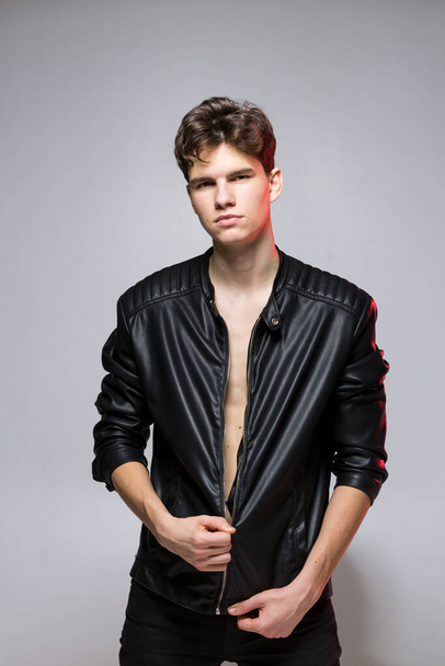 Handsome Athletic Male Model Wearing Leather Jacket on Naked Torso. Sexy young guy with a beautiful body in a biker jacket on a red background in the studio. Fashionable young Man in leather coat. - Zdjęcie, obraz