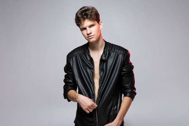 Handsome caucasian guy model with naked tors, lack leather jacket posing in studio on red background. Sexy young man in black stylish clothes on naked body. Emotional posing, lifestyle people concept. - Photo, Image