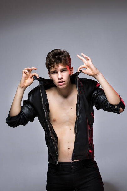 Handsome Athletic Male Model Wearing Leather Jacket on Naked Torso. Sexy young guy with a beautiful body in a biker jacket on a red background in the studio. Fashionable young Man in leather coat. - Photo, Image