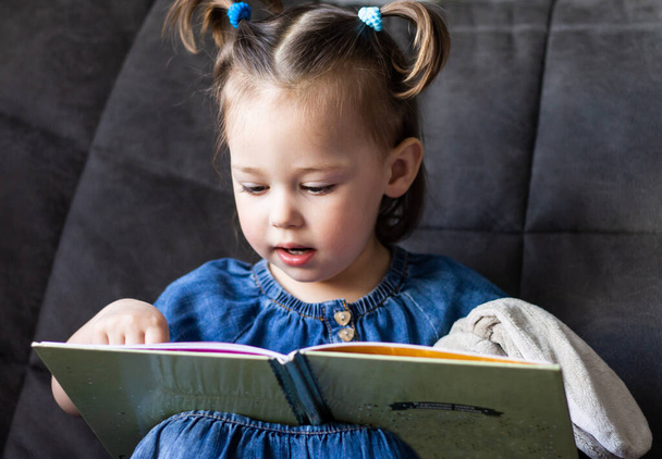 Lapsen lukemista. Pieni söpö tyttö 1-3 lukee kirjaa, jossa lelu plush jänis istuu sohvalla - Valokuva, kuva
