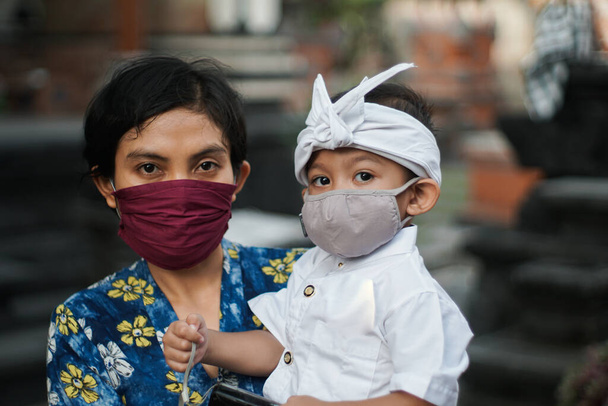 Балийская мать и ребенок одеты в традиционную одежду во время пандемии короны или ковида-19. Они оба используют маски, чтобы защитить себя от вирусных атак. Они готовятся молиться в храме - Фото, изображение