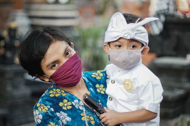 Madre e figlio balinesi indossano abiti tradizionali durante la pandemia della corona o covid-19. Entrambi usano maschere per proteggersi dagli attacchi di virus. Si preparano a pregare al tempio - Foto, immagini