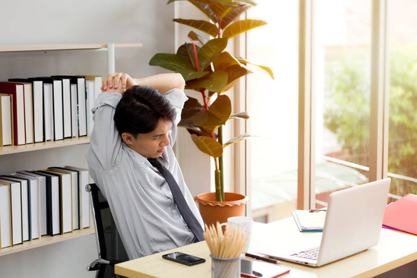 Asiatischer männlicher Angestellter saß an einem Schreibtisch, er fühlt sich müde von der Arbeit und hebt die Arme auf den Kopf, um sich zu strecken. - Foto, Bild