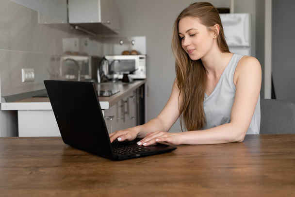 Портрет молодой симпатичной белой женщины, работающей или обучающейся дистанционно, с помощью ноутбука на кухне. Изображение в помещении, технологии, онлайн концепция - Фото, изображение