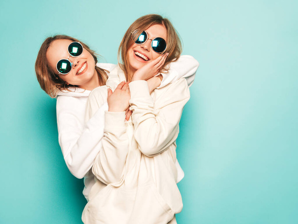 Two young beautiful blond smiling hipster girls in trendy summer hoodie clothes. Sexy carefree women posing near blue wall. Trendy and positive models having fun in sunglasses - Foto, Imagem