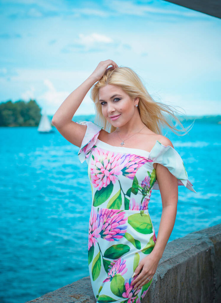 Beautiful blonde hair woman in romantic flowers dress near blue water and sand. Cute happy girl have fun on a nature in vacation - Fotó, kép