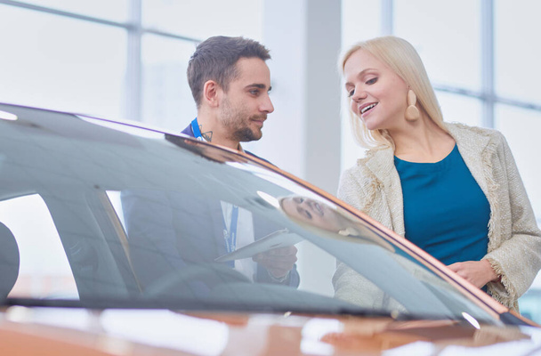 Dealer with woman stands near a new car in the showroom - Foto, Imagem