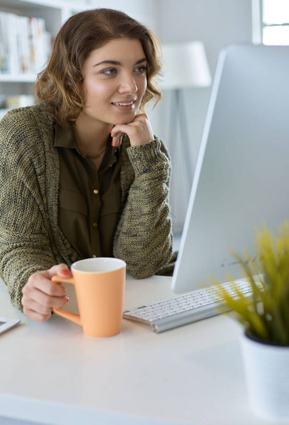 Attractive smiling woman sitting at office desk, holding a cup of coffee, she is relaxing and looking away - Foto, Bild